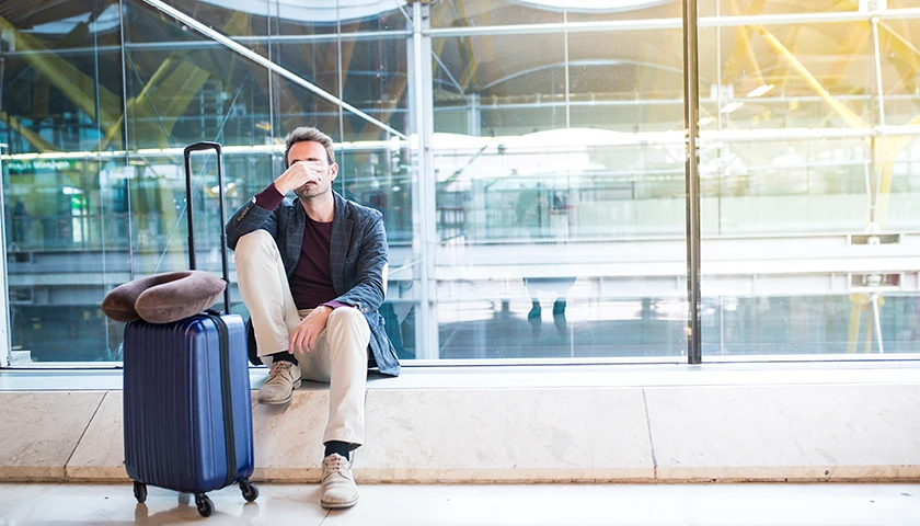 Hombre cansado en el aeropuerto, sentado junto a su maleta de mano, pensando en tomar melatonina para aliviar el jet lag.webp