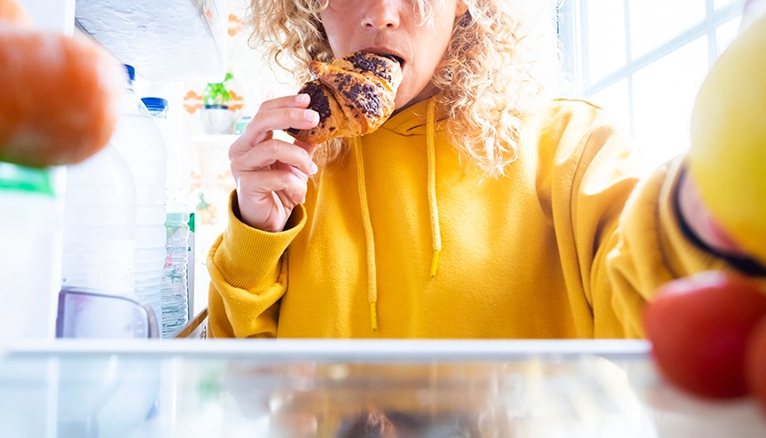 Persona con hambre emocional comiendo un bollo delante de la nevera abierta.webp